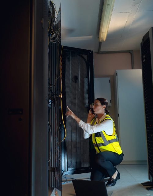Caucasian female engineer with laptop talking on smartphone while inspecting in computer server room. database server management and maintenance concept