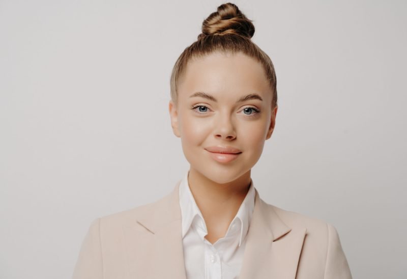 Close up portrait of young pretty brunette business lady with hair bun wearing formal clothes with white shirt standing isolated in front of grey background looking forward with confidence
