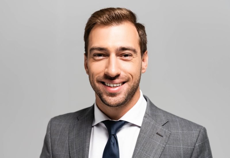 handsome and smiling businessman in suit looking at camera isolated on grey
